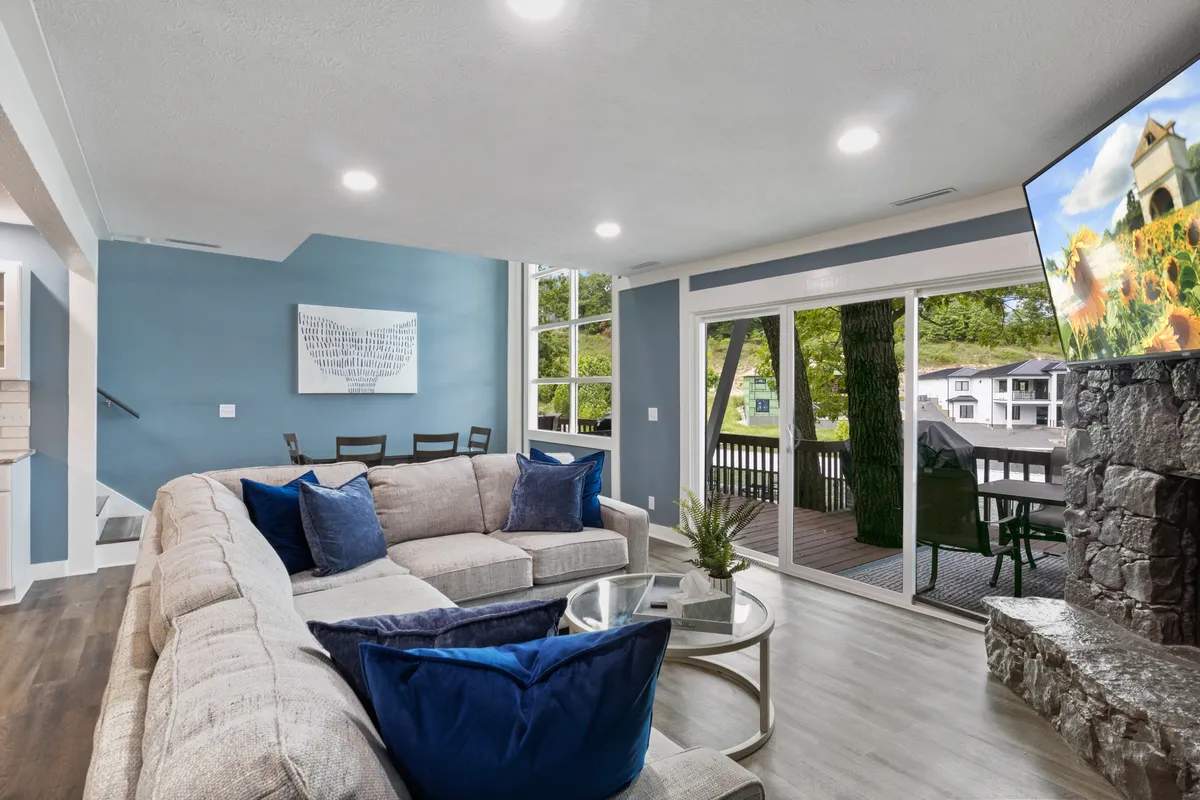 This image presents a spacious living room featuring a large sectional sofa, a glass coffee table, a wall-mounted TV, and expansive windows that look out onto a deck. The room feels open, cozy, and is flooded with natural light, perfectly capturing areas for family gatherings, relaxing, or watching movies. The view and deck access reinforce the connection between the interior and the lakeside setting.