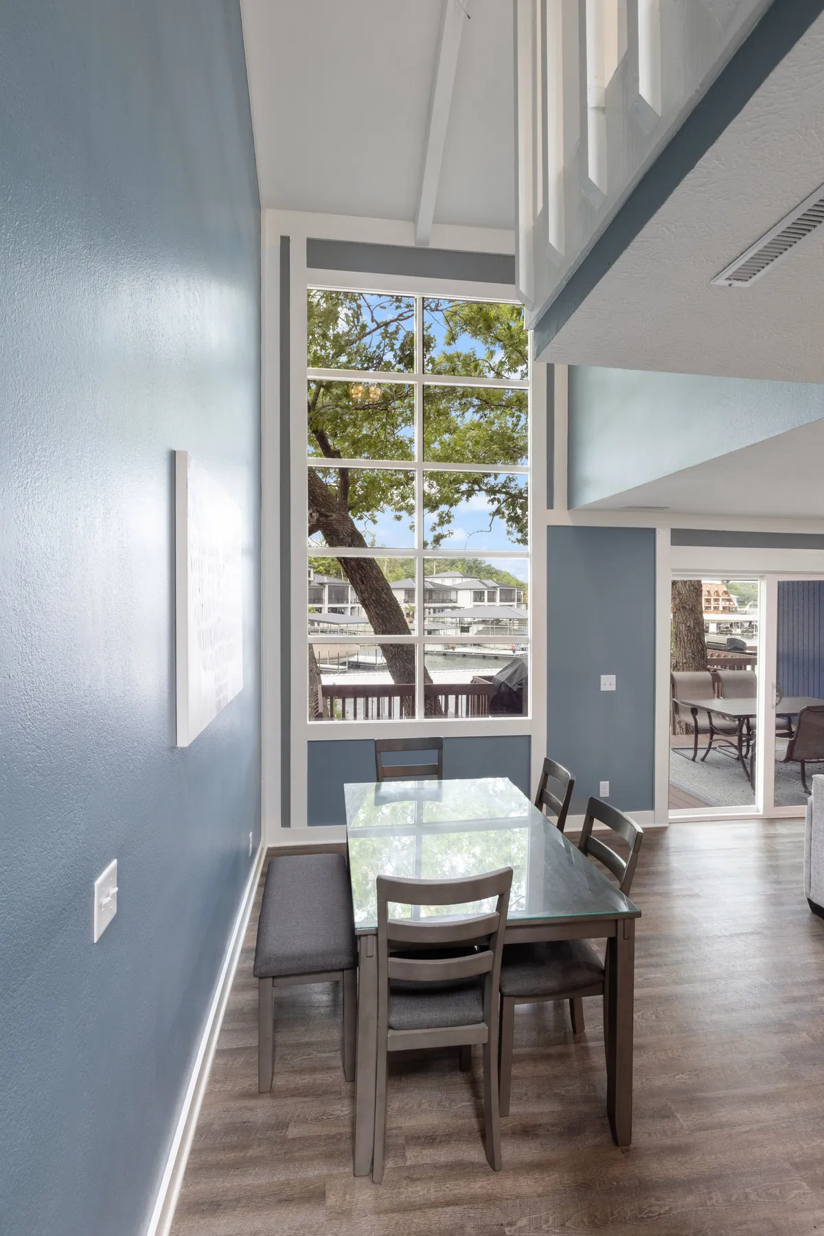 A sunlit dining area features a large table set before wide windows, allowing generous sunlight and views of the outdoors to pour in. The scene suggests a space perfect for shared meals, celebrations, or afternoon relaxation, with the flooring and windows visually linking to adjoining living areas.