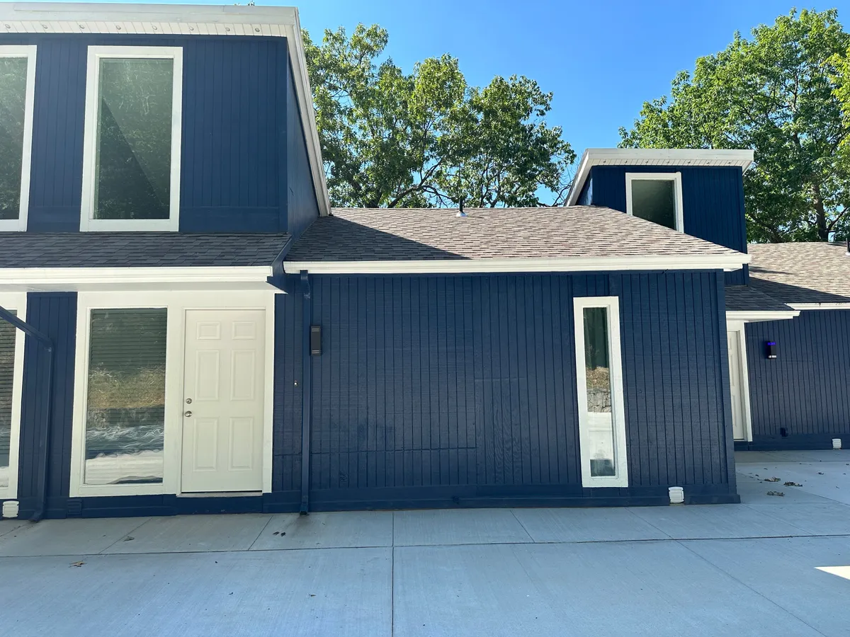 A modern exterior view of the property, featuring dark blue siding, expansive windows, and a welcoming white door. The porch area hints at possibilities for outdoor relaxation and gatherings, with the architecture opening toward the surrounding environment.
