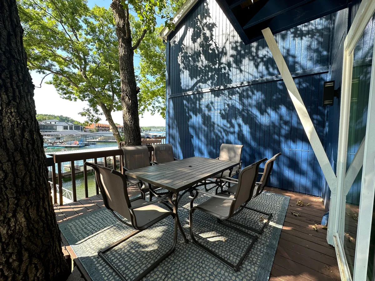 An inviting outdoor dining space is set among trees with a clear view of the water, offering a tranquil environment for outdoor meals. The patio table and surrounding greenery suggest a seamless transition between indoor comfort and the refreshing lakeside outdoors.