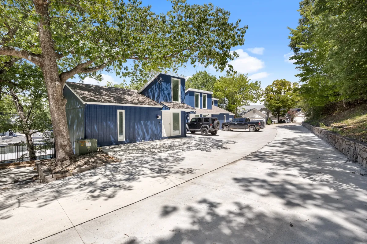 This image showcases the blue exterior of the house surrounded by mature trees, with a welcoming parking area and driveway. There is ample open space suggesting room for kids to play and for families to gather outdoors. The inviting façade, shaded areas, and visible access to outdoor living spaces set an approachable, family-friendly atmosphere.
