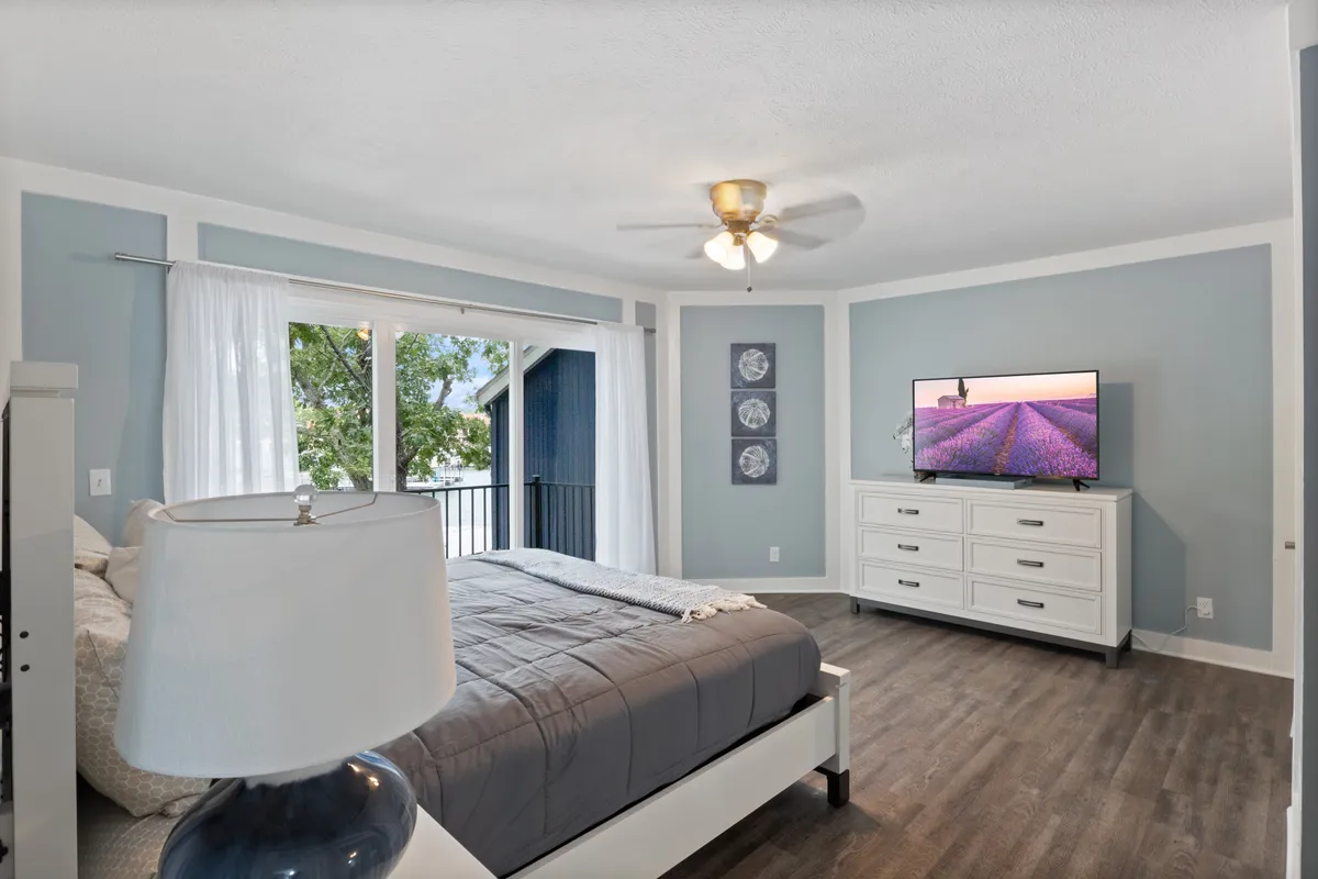 A serene bedroom with gray bedding and a white dresser topped with a TV, framed by large windows dressed in sheer curtains. Outside, a balcony is visible, hinting at inviting sunset or sunrise views. The atmosphere is peaceful and inviting, perfect for unwinding.
