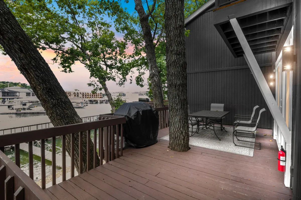 The shot captures an outdoor dining set and grill positioned on a deck or patio overlooking the water, framed by leafy trees. The tranquil water view and natural surroundings suggest a serene lakeside experience, with spaces for adults to relax and keep an eye on children near the water. The scene evokes barbecues, picnics, and relaxed supervision of lakeside activities.