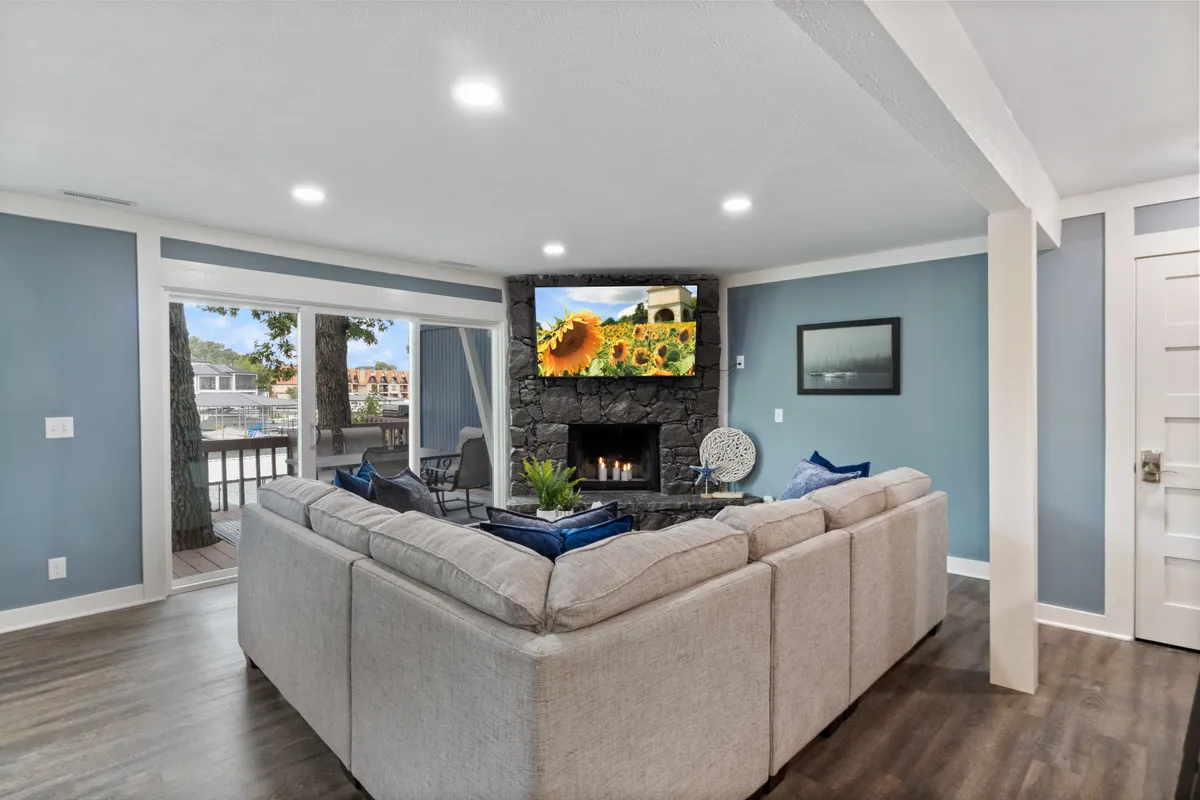 This inviting interior features a spacious sectional sofa centered around a cozy fireplace and a large TV, all set against a backdrop of expansive windows that offer lake views and glowing sunsets. The warm, welcoming atmosphere is perfect for family gatherings, movie nights, or simply relaxing while soaking in the serene surroundings.