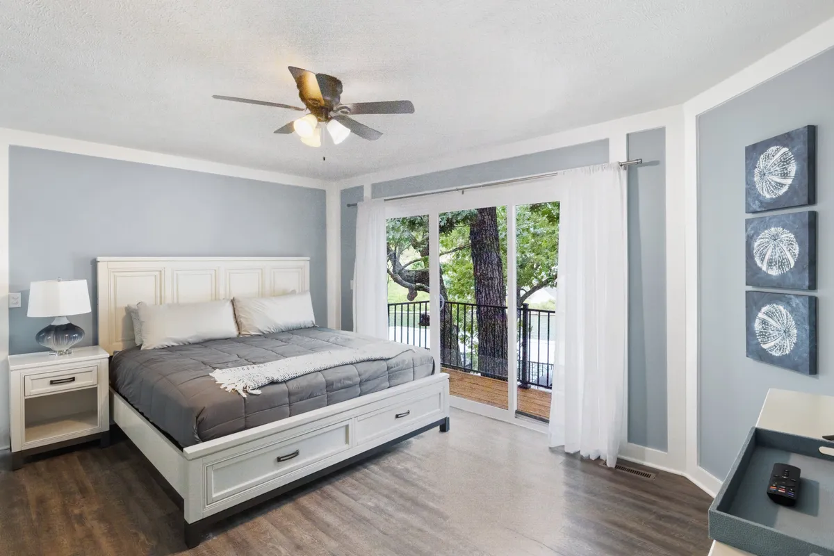 A tranquil bedroom scene featuring a bed positioned beside a sliding glass door, with a nightstand completing the cozy arrangement. Soft morning light gently filters through sheer curtains, and the door appears to open onto a private balcony or deck, suggesting easy access to outdoor relaxation.