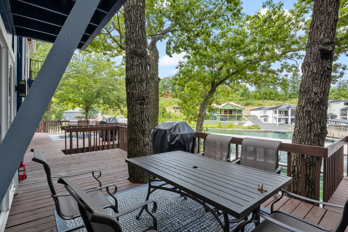 An inviting outdoor dining area with a dining table set under the shade of mature trees, offering a picturesque view of the water. A barbecue grill stands nearby, suggesting opportunities for al fresco meals while enjoying the lake breeze. The scene exudes relaxation and a connection to nature, perfect for spending time with family and pets after a lakeside stroll.